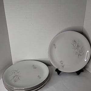 May include: A set of four round white ceramic plates featuring a delicate grey line drawing of a rose and its stem. The plates have a smooth finish and a thin metallic silver rim around the edge. One plate is displayed upright on a black stand.