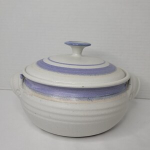 May include: A round ceramic casserole dish with a matching lid, featuring a speckled off-white base and horizontal bands of soft lavender purple. The pot includes two side handles and a small knob on the lid, with a textured, ribbed exterior.