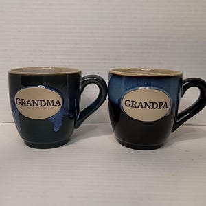 May include: Two ceramic mugs with a dark blue and black reactive glaze finish. Each mug features a central oval-shaped beige medallion. The left mug has the word GRANDMA printed in black, and the right mug has the word GRANDPA printed in black.