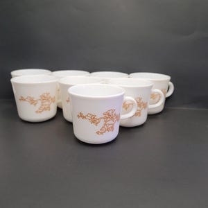 May include: A collection of white ceramic mugs with a brown floral design. Each mug has a handle and a slightly flared shape. The mugs are arranged on a dark surface, showcasing their simple elegance.