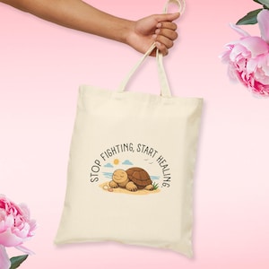 May include: Cream-colored tote bag with a cartoon turtle illustration. The turtle is lying on a beach with the text "Stop Fighting, Start Healing" in a circular design. The bag is held by a hand against a pink background.
