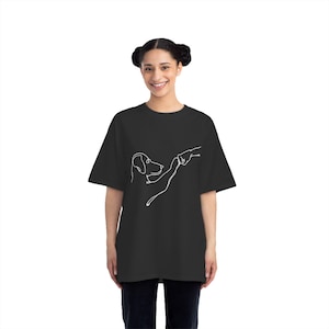 Op de afbeelding: Zwart T-shirt met een witte lijntekening van een hond die een menselijke hand aanraakt. Het ontwerp staat in het midden. Het shirt heeft korte mouwen en een losse pasvorm.