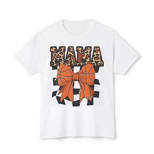 May include: White t-shirt with a basketball-themed design. The word "MAMA" is in a leopard print font above a large orange bow with basketball textures. A black and white checkered pattern forms the background of the bow.