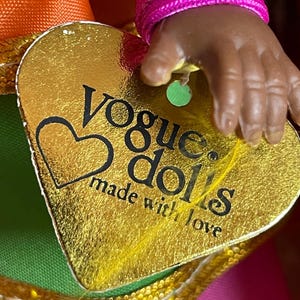 May include: A gold heart-shaped tag with the words "Vogue dolls made with love" is held by a doll's hand. The tag is attached to a doll's outfit with orange, green, and pink fabric.