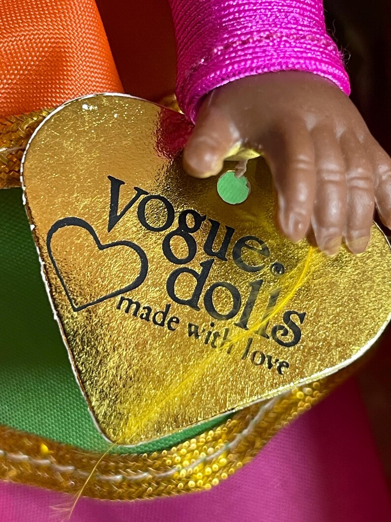 May include: A gold heart-shaped tag with the words "Vogue dolls made with love" is held by a doll's hand. The tag is attached to a doll's outfit with orange, green, and pink fabric.