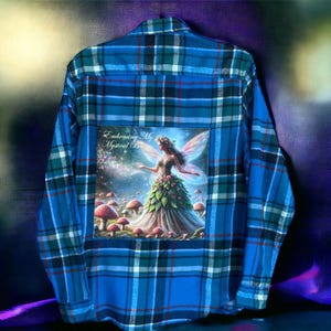 May include: A blue plaid flannel shirt with a patch on the back featuring a fairy with wings, surrounded by mushrooms and the text "Embracing My Mystical Beauty".