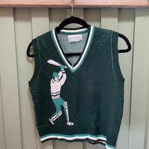 May include: A dark green V-neck sweater vest featuring a white and light green intarsia graphic of a cricket player swinging a bat. The garment has white and light green striped trim at the neckline and hem, with a Bobbie Brooks brand label inside.
