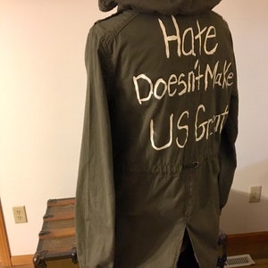 May include: A green hooded jacket with a white message on the back that reads "Hate Doesn't Make Us Great".