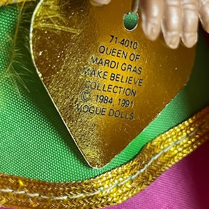 May include: A close-up of a gold heart-shaped tag with the text "QUEEN OF MARDI GRAS MAKE BELIEVE COLLECTION" and the Vogue Dolls logo. The tag is attached to a doll's outfit with pink, green, and gold accents. The doll's hand is visible.