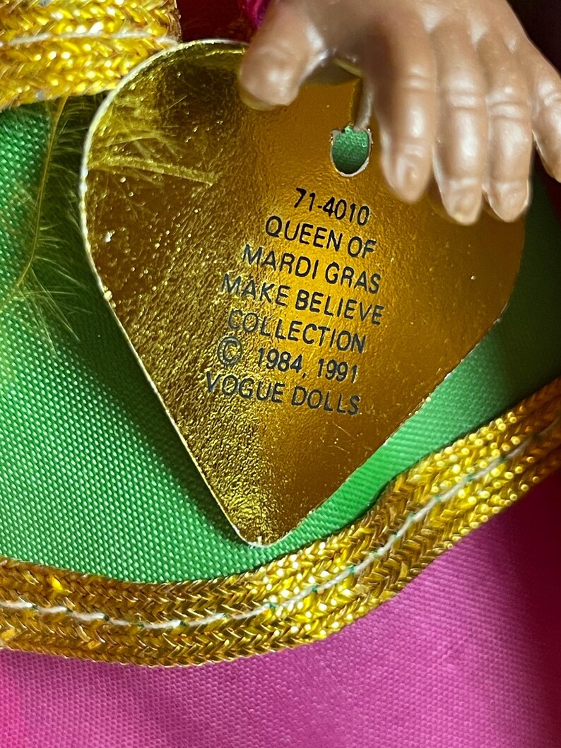 May include: A close-up of a gold heart-shaped tag with the text "QUEEN OF MARDI GRAS MAKE BELIEVE COLLECTION" and the Vogue Dolls logo. The tag is attached to a doll's outfit with pink, green, and gold accents. The doll's hand is visible.