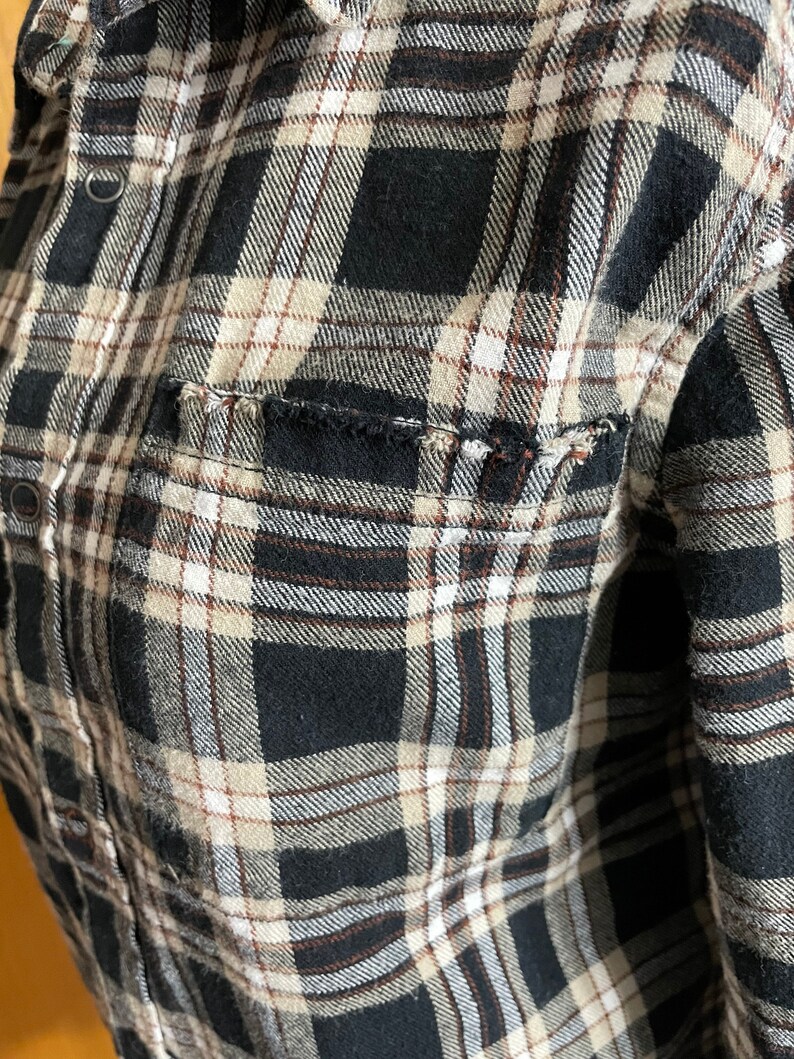 May include: A black and white plaid flannel shirt with brown accents. The shirt has a pocket on the left side and a frayed edge.