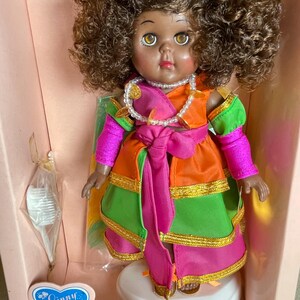 May include: A Ginny doll, the "Queen of Mardi Gras," in a pink box. The doll has brown curly hair, brown eyes, and is wearing a colorful costume with orange, green, and pink accents. A pearl necklace and gold trim complete the look.