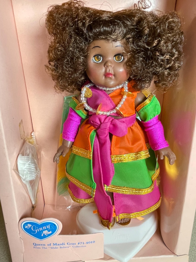 May include: A Ginny doll, the "Queen of Mardi Gras," in a pink box. The doll has brown curly hair, brown eyes, and is wearing a colorful costume with orange, green, and pink accents. A pearl necklace and gold trim complete the look.