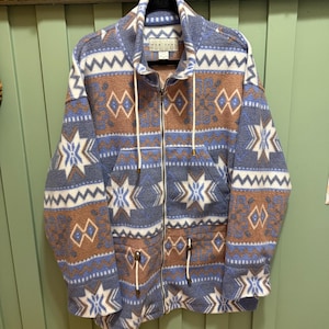 May include: A zip-up fleece jacket featuring a blue, tan, and white geometric pattern with star and diamond motifs. The jacket has a high collar, long sleeves, two front pockets, and white drawstring cords with metal tips at the neck and waist.