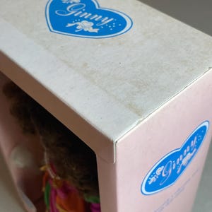 May include: A close-up of a doll box with the name "Ginny" in a blue heart-shaped logo. The box is pink and white, with the doll's hair and clothing visible through the opening. The box is slightly worn.