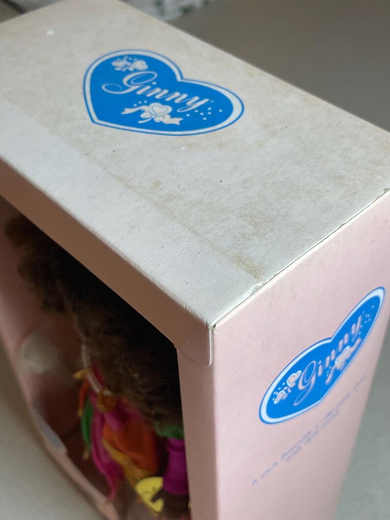 May include: A close-up of a doll box with the name "Ginny" in a blue heart-shaped logo. The box is pink and white, with the doll's hair and clothing visible through the opening. The box is slightly worn.