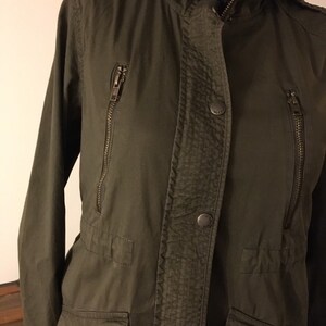 May include: A green hooded jacket with a zipper closure and multiple pockets. The jacket has a buttoned flap over the zipper.