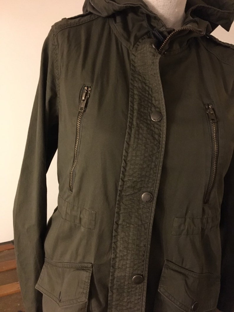 May include: A green hooded jacket with a zipper closure and multiple pockets. The jacket has a buttoned flap over the zipper.