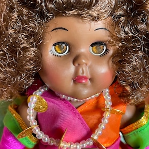 May include: A vintage doll with brown curly hair and large, expressive eyes. The doll wears a colorful outfit with pink, orange, and green accents, and a beaded necklace with gold-colored embellishments. The doll's face has a sweet, innocent expression.