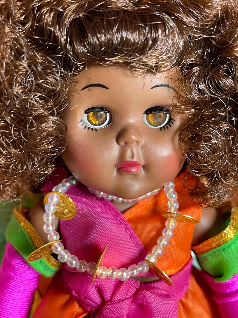 May include: A vintage doll with brown curly hair and large, expressive eyes. The doll wears a colorful outfit with pink, orange, and green accents, and a beaded necklace with gold-colored embellishments. The doll's face has a sweet, innocent expression.