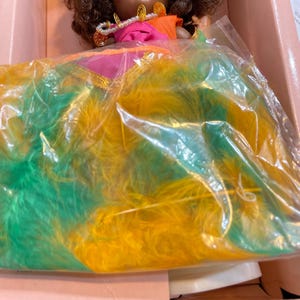 May include: A vintage Vogue doll in its original box. The doll has brown curly hair, a pink and orange top, and a necklace. The doll is wearing a dress with green and yellow feathers. The box is pink and has the Vogue Dolls logo.