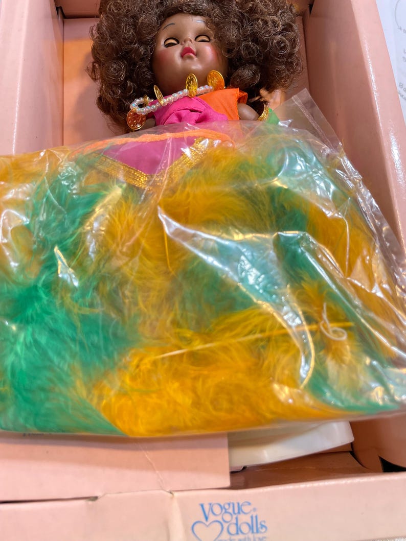 May include: A vintage Vogue doll in its original box. The doll has brown curly hair, a pink and orange top, and a necklace. The doll is wearing a dress with green and yellow feathers. The box is pink and has the Vogue Dolls logo.
