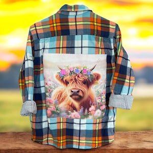 May include: A blue, orange, and white plaid flannel shirt with a printed image of a Highland cow wearing a flower crown. The cow is brown and white and has a soft, fuzzy coat.