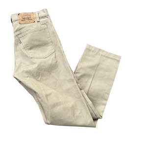 May include: Beige Levi Strauss & Co. pants featuring a tan leather patch on the back waistband, belt loops, and a single back pocket with a small white tab. The pants are constructed from a solid-colored cotton twill fabric with straight legs.