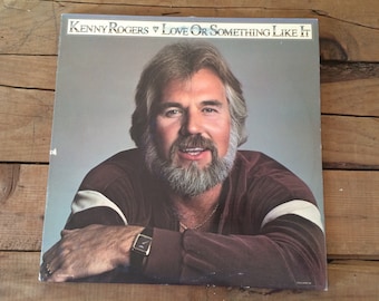 May include: A vinyl record album cover featuring Kenny Rogers with a beard and mustache. The album title is "Love Or Something Like It".
