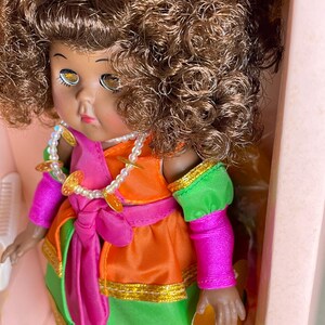 May include: A vintage doll with brown curly hair, wearing a colorful outfit. The doll's dress is a combination of orange, green, and pink, with gold trim and a matching pink sash. A pearl necklace with gold coin accents adorns the doll's neck.