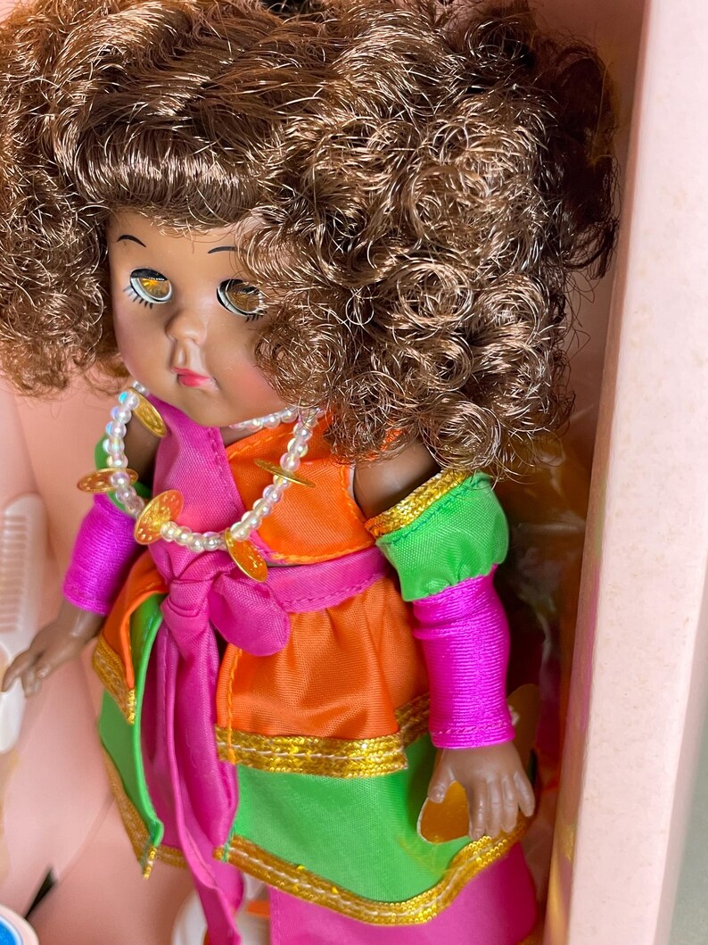May include: A vintage doll with brown curly hair, wearing a colorful outfit. The doll's dress is a combination of orange, green, and pink, with gold trim and a matching pink sash. A pearl necklace with gold coin accents adorns the doll's neck.