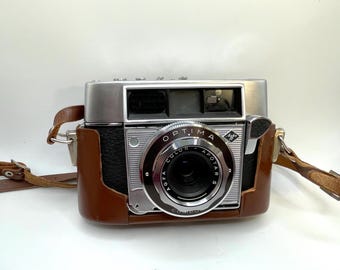 May include: A vintage Agfa Optima Color Apotar S camera in a brown leather case. The camera has a silver top and a black body with a prominent lens. The lens is surrounded by a silver ring with the text "Optima Color Apotar S". A brown leather strap is attached.