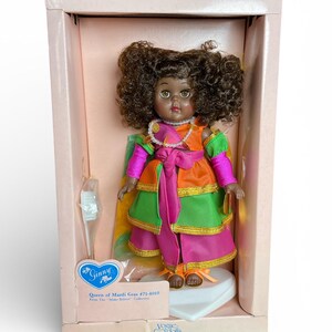 May include: A vintage Ginny doll, "Queen of Mardi Gras #71-4010," in its original box. The doll has brown skin, curly brown hair, and is dressed in a colorful layered gown with pink, green, and orange accents. The box is light pink.