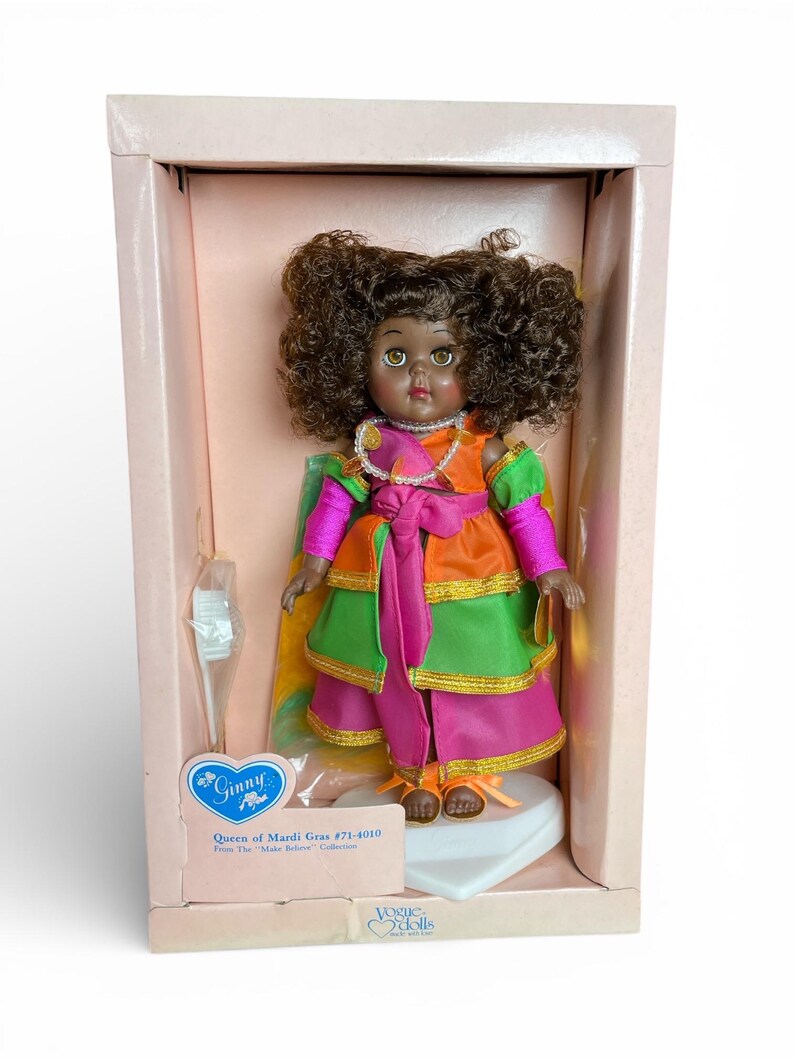 May include: A vintage Ginny doll, "Queen of Mardi Gras #71-4010," in its original box. The doll has brown skin, curly brown hair, and is dressed in a colorful layered gown with pink, green, and orange accents. The box is light pink.