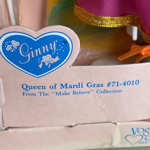May include: A vintage Ginny doll box featuring the text "Ginny" in a blue heart-shaped logo. The box reads "Queen of Mardi Gras #71-4010" from the "Make Believe" Collection. The doll's colorful dress is visible.