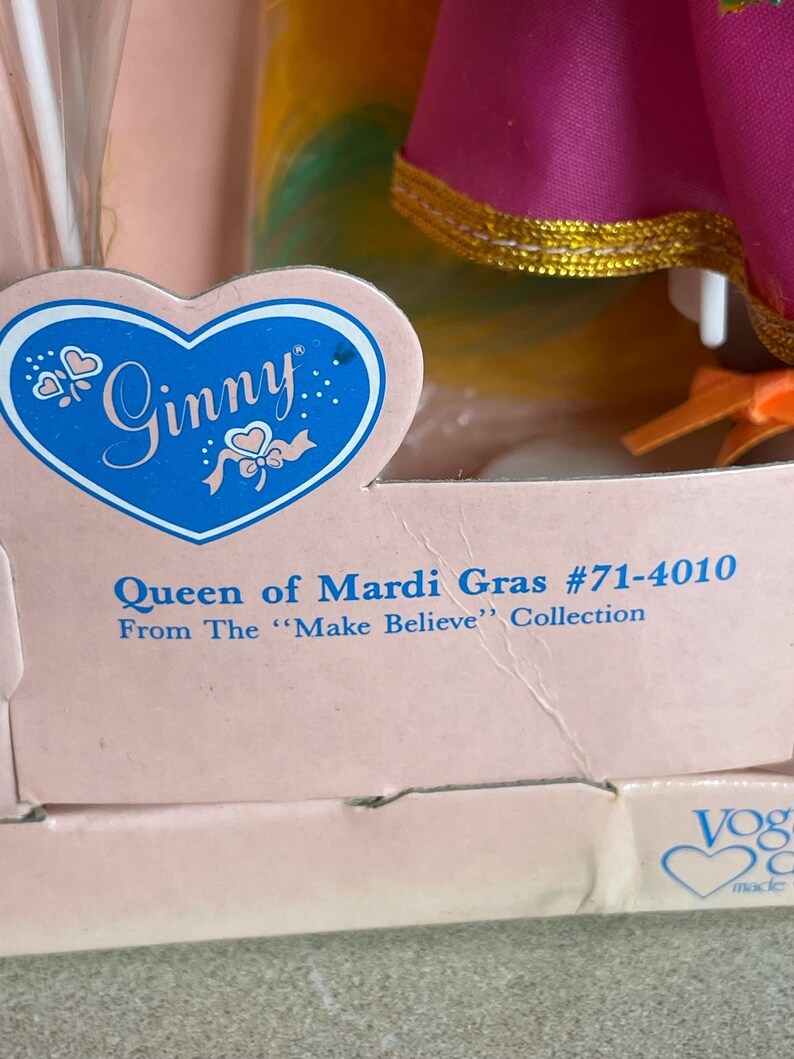 May include: A vintage Ginny doll box featuring the text "Ginny" in a blue heart-shaped logo. The box reads "Queen of Mardi Gras #71-4010" from the "Make Believe" Collection. The doll's colorful dress is visible.
