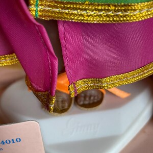May include: Close-up of a doll wearing a green and fuchsia outfit with gold trim. The doll's outfit has a gold trim. The doll is standing on a white heart-shaped base with the name "Ginny" embossed on it.