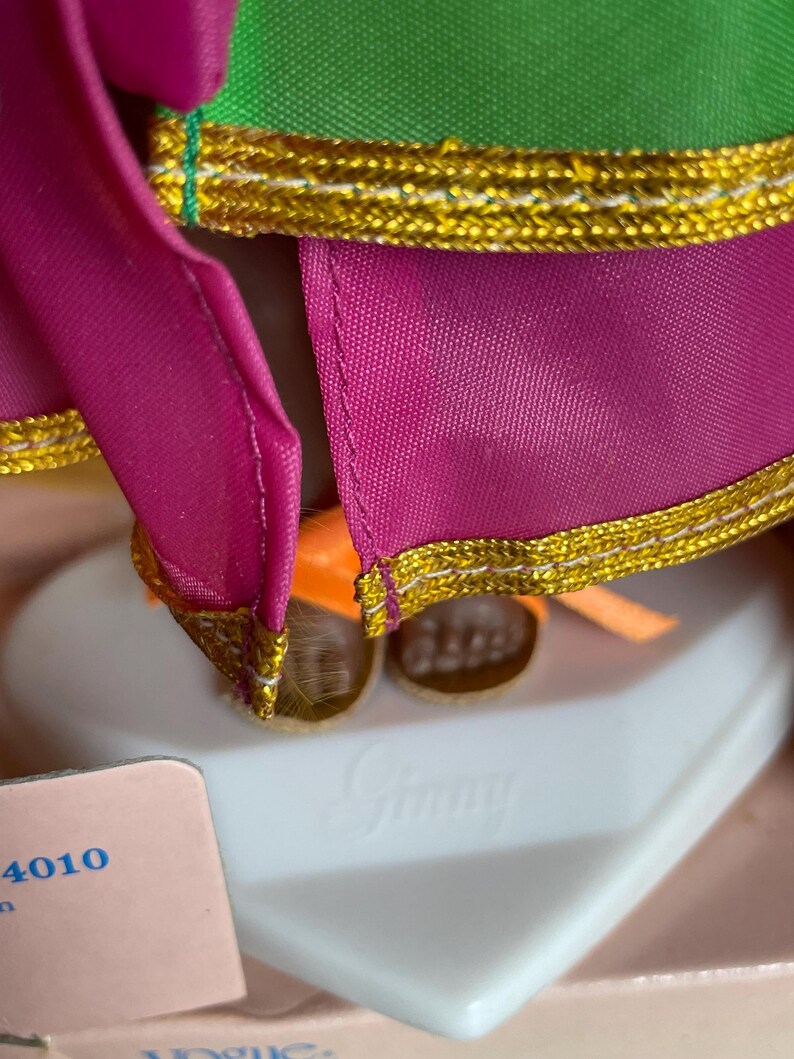 May include: Close-up of a doll wearing a green and fuchsia outfit with gold trim. The doll's outfit has a gold trim. The doll is standing on a white heart-shaped base with the name "Ginny" embossed on it.