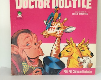 May include: A pink vinyl record album cover featuring the title "Doctor Dolittle" in large white letters. The cover art includes a cartoon monkey holding a top hat, a giraffe with a parrot on its head, and two dogs. The text "Music and Lyrics by Leslie Bricusse" is printed in white on the right side of the cover.