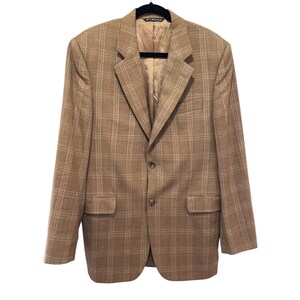May include: A tan plaid blazer with a two-button closure, notched lapels, and two front pockets. The blazer is hanging on a black hanger against a white background. The interior lining is a solid tan color.