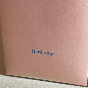 May include: A section of a light pink box with the words "Hard vinyl" printed in blue. The box has a matte finish and is sitting on a light-colored surface.
