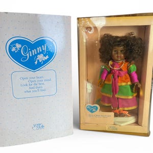 May include: A Ginny doll in a box. The doll has dark skin, curly hair, and is wearing a colorful dress with pink, orange, and green accents. The box has the Ginny logo and the text "Open your heart, Open your mind, Look for the best, And that's what you'll find."