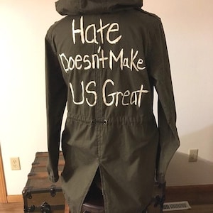 May include: A green hooded jacket with a white message on the back that reads "Hate Doesn't Make Us Great".