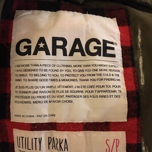 May include: A green utility parka with a red and black plaid lining. The label reads "Garage" and includes the text "I am more than a piece of clothing, more than you might expect. I was designed to be found by you. To give you one more reason to smile. To belong to you. To protect you from the cold & the wind. To share good times & memories. Thank you for finding me."  The label also includes the text "Utility Parka" and "Trust your instinct - Fie toi &agrave; ton instinct". The size tag reads "S/P".