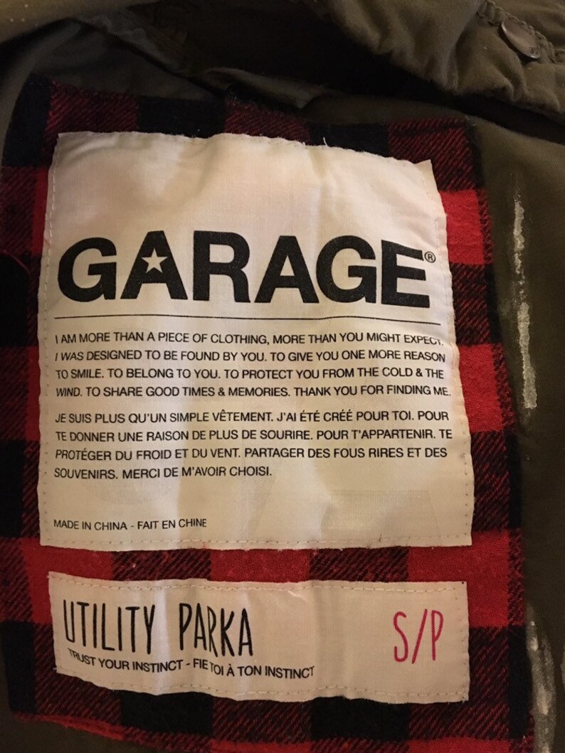 May include: A green utility parka with a red and black plaid lining. The label reads "Garage" and includes the text "I am more than a piece of clothing, more than you might expect. I was designed to be found by you. To give you one more reason to smile. To belong to you. To protect you from the cold & the wind. To share good times & memories. Thank you for finding me."  The label also includes the text "Utility Parka" and "Trust your instinct - Fie toi &agrave; ton instinct". The size tag reads "S/P".