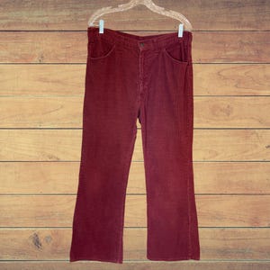 May include: A pair of deep maroon corduroy pants featuring a high-waisted design, a button and zipper fly closure, two front pockets, and a straight-leg cut. The pants are displayed on a clear plastic hanger against a horizontal wood plank background.