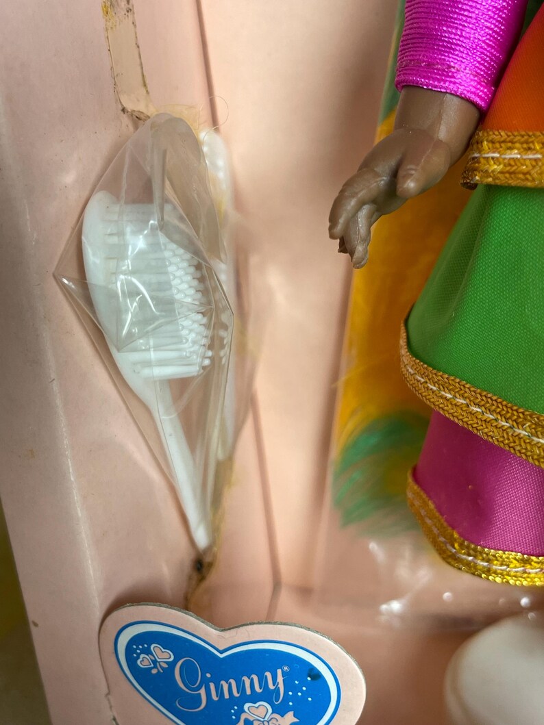 May include: A vintage Ginny doll in a pink box. The doll has a colorful dress with gold trim and a pink top. A white toothbrush is in a plastic bag. The Ginny label is visible.