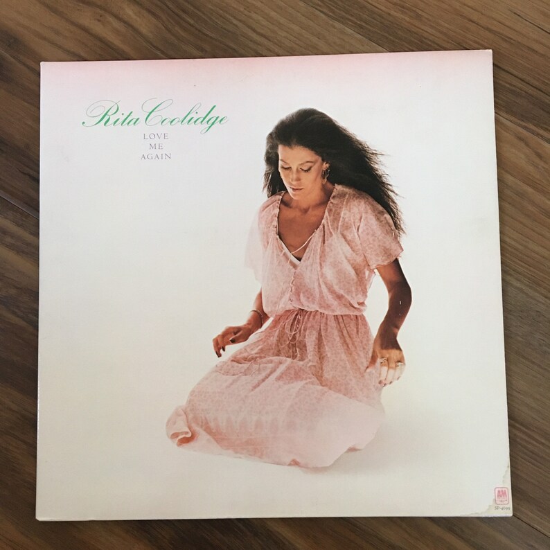 May include: A vintage vinyl record album cover featuring Rita Coolidge. The cover is white with a pink background and features a photo of Rita Coolidge wearing a pink dress. The album title is "Love Me Again" and is printed in green text.
