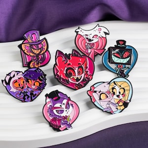 Puede incluir: Una colección de coloridos pines de esmalte con varios personajes de dibujos animados. Los pines muestran una variedad de diseños, incluyendo pines en forma de corazón y de personajes en tonos de rosa, morado, rojo y azul. El fondo es una superficie blanca ondulada.