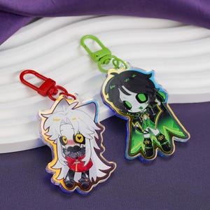 Puede incluir: Dos llaveros de acrílico con personajes de estilo anime. Un llavero tiene un cierre rojo y un personaje con cabello blanco y traje rojo. El otro tiene un cierre verde y un personaje con cabello negro y traje verde.
